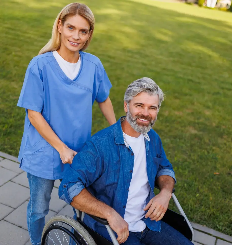 A woman in medical scrubs stands behind a smiling man seated in a wheelchair outdoors on a paved path with grass in the background.