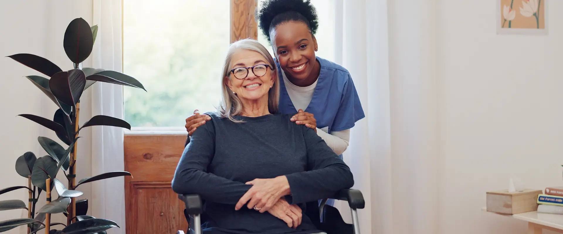 A smiling elderly woman in a wheelchair sits by a window while a healthcare worker in scrubs stands behind her, both looking at the camera.