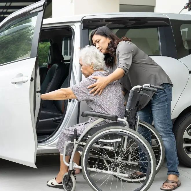 A woman assists an elderly person in a wheelchair with getting into a car, holding her arm for support.