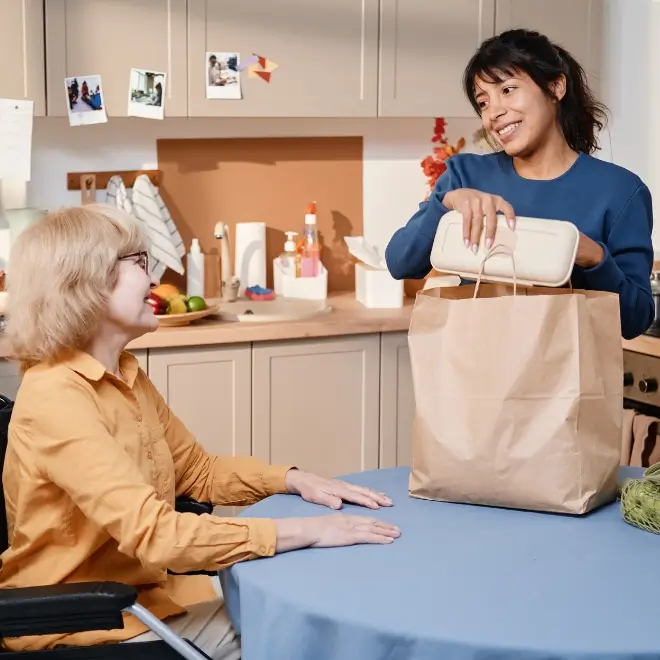 A woman sits at a table in a wheelchair while another woman hands her a food container from a paper bag in a kitchen.