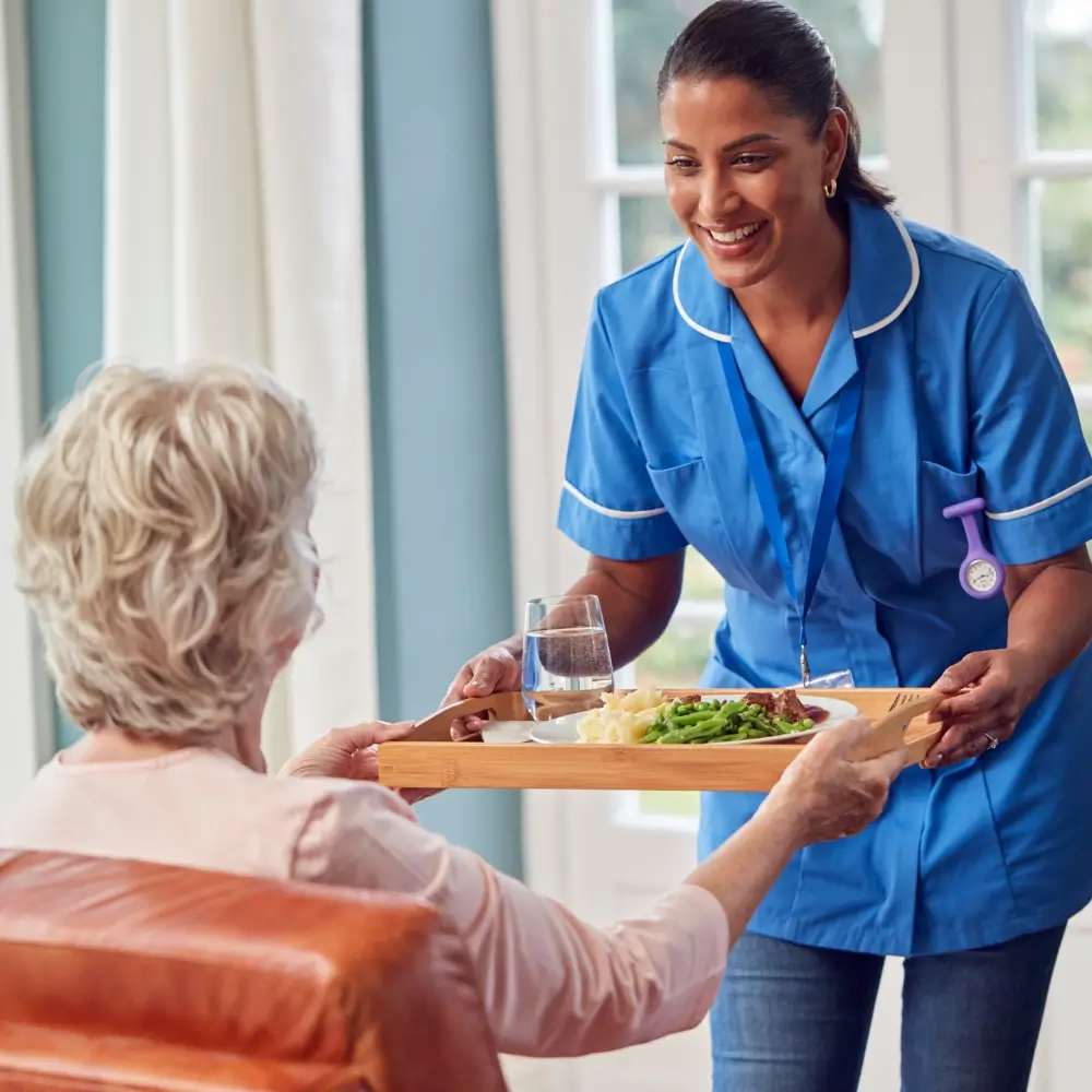 A caregiver in a blue uniform hands a tray with a meal and a glass of water to an elderly woman seated in a living room.