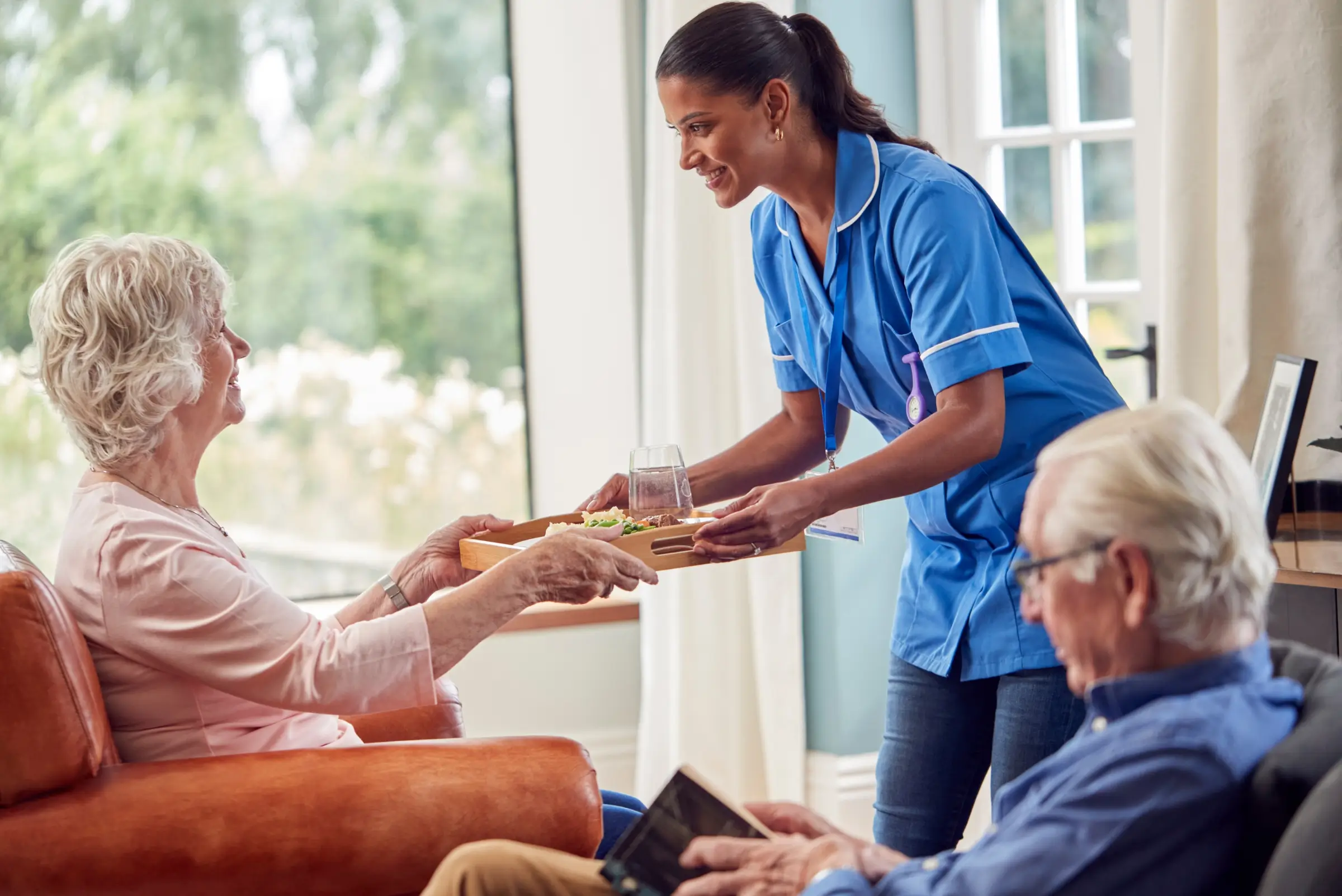 A caregiver in uniform hands a meal tray to an elderly woman sitting in a chair, while an elderly man reads beside her, reflecting quality domestic assistance in a bright living room.