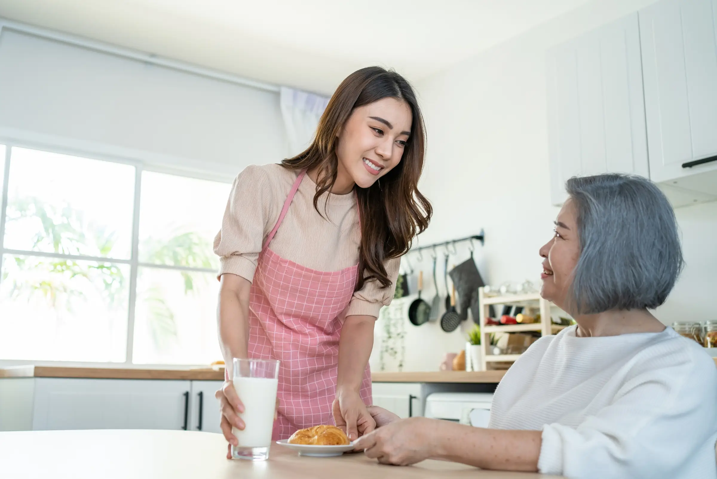 A young woman in an apron provides quality home care, serving a glass of milk and a plate of pastries to an older woman sitting at a kitchen table.