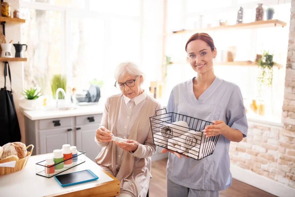 An elderly woman sits at a kitchen table with medicine bottles, while a caregiver in scrubs—providing domestic assistance—stands nearby holding a basket of folded towels, supporting her wellbeing and independence.