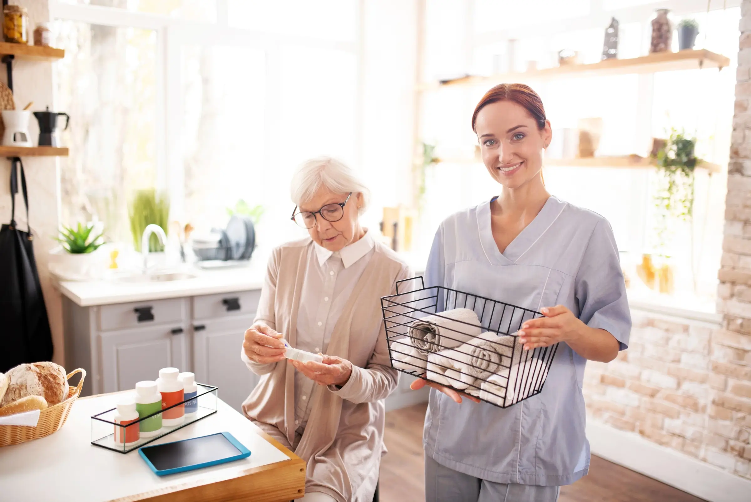 An elderly woman sits at a kitchen table with medicine bottles, while a caregiver in scrubs—providing domestic assistance—stands nearby holding a basket of folded towels, supporting her wellbeing and independence.