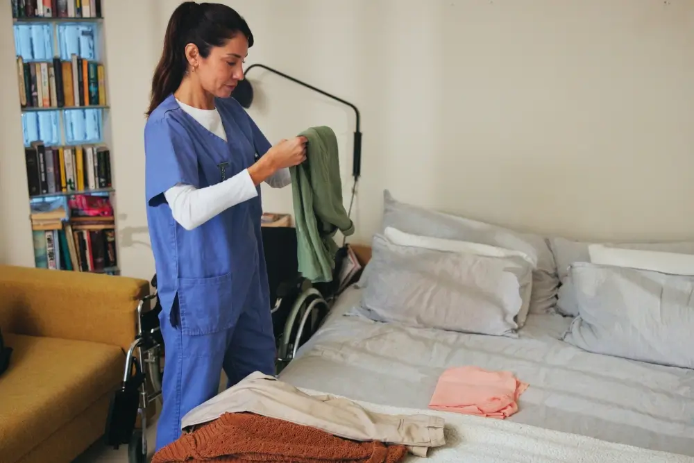 A woman in scrubs stands by a bed, folding laundry in a bedroom with a wheelchair, bookshelf, and sofa visible.