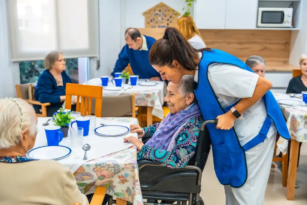 A caregiver in a blue vest speaks to a smiling elderly woman in a wheelchair at a dining table in a communal care facility. Other elderly people are seated at nearby tables.