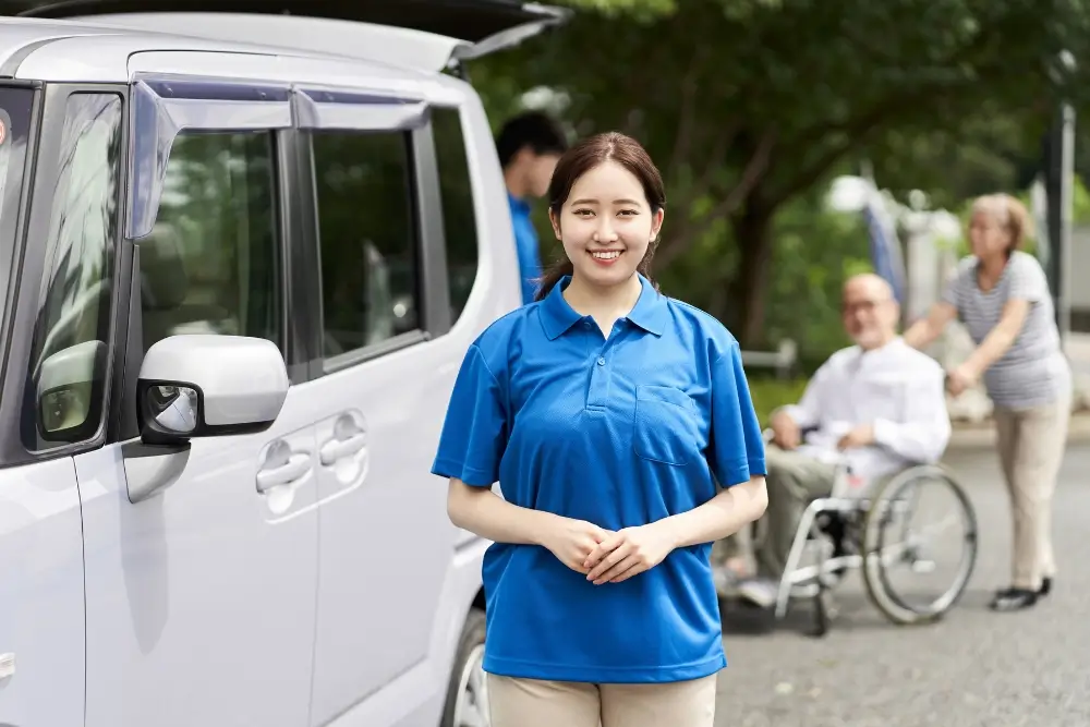 A woman in a blue polo shirt stands by a white vehicle, while a person in a wheelchair is pushed by another person in the background.