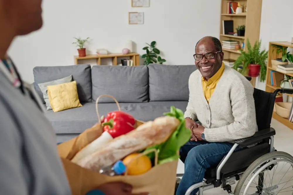 A man in a wheelchair smiles at a person holding a grocery bag filled with fresh produce and bread in a living room.