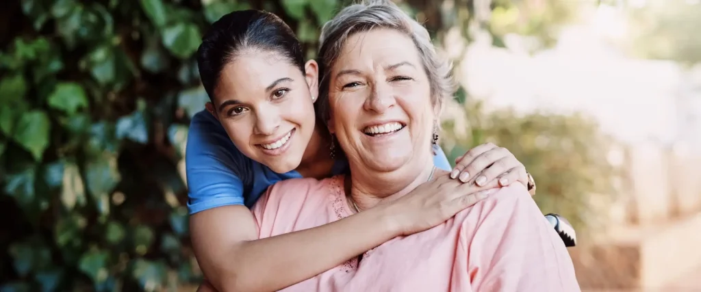 A younger woman in blue scrubs stands behind an older woman in pink, smiling and hugging her shoulders. They are outdoors with greenery in the background.