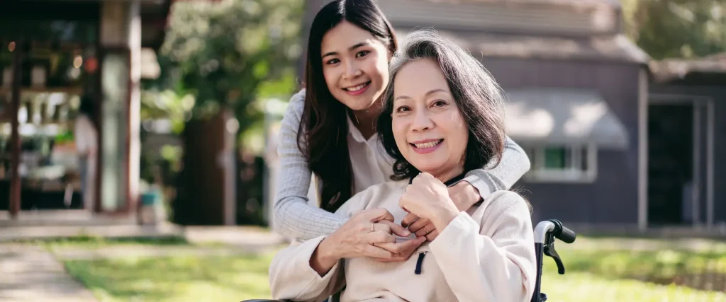A smiling older woman sits in a wheelchair while a younger woman stands behind her, embracing her shoulders, outdoors in a garden setting.