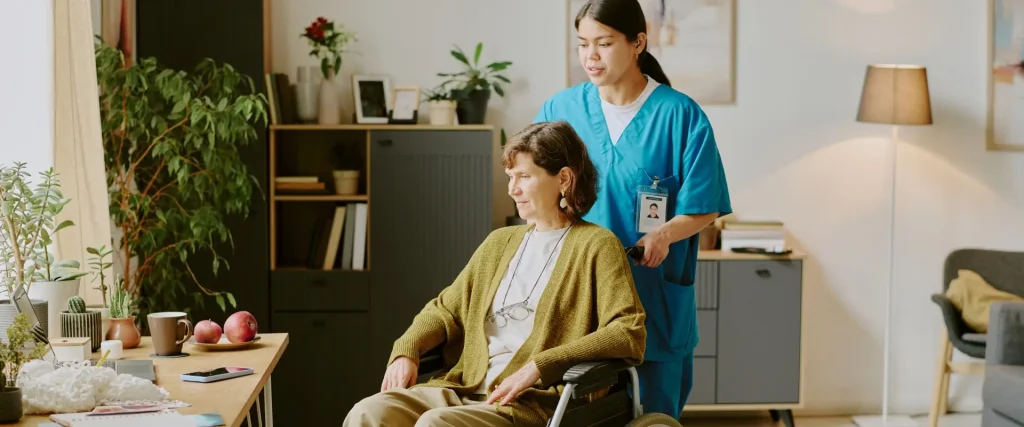 A healthcare worker in scrubs stands behind an older woman sitting in a wheelchair in a well-lit living room with plants and bookshelves.