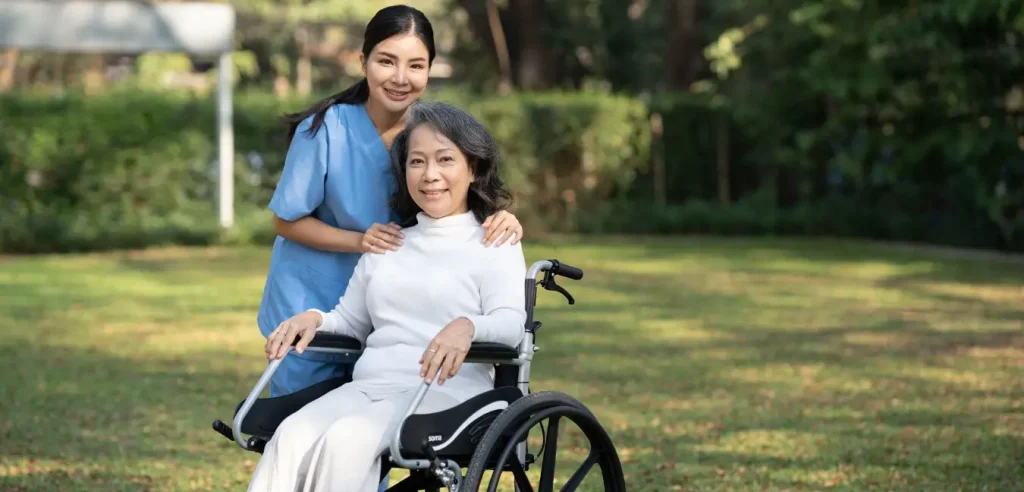 A caregiver stands behind an older woman in a wheelchair, both smiling, outdoors on a grassy area with trees in the background.